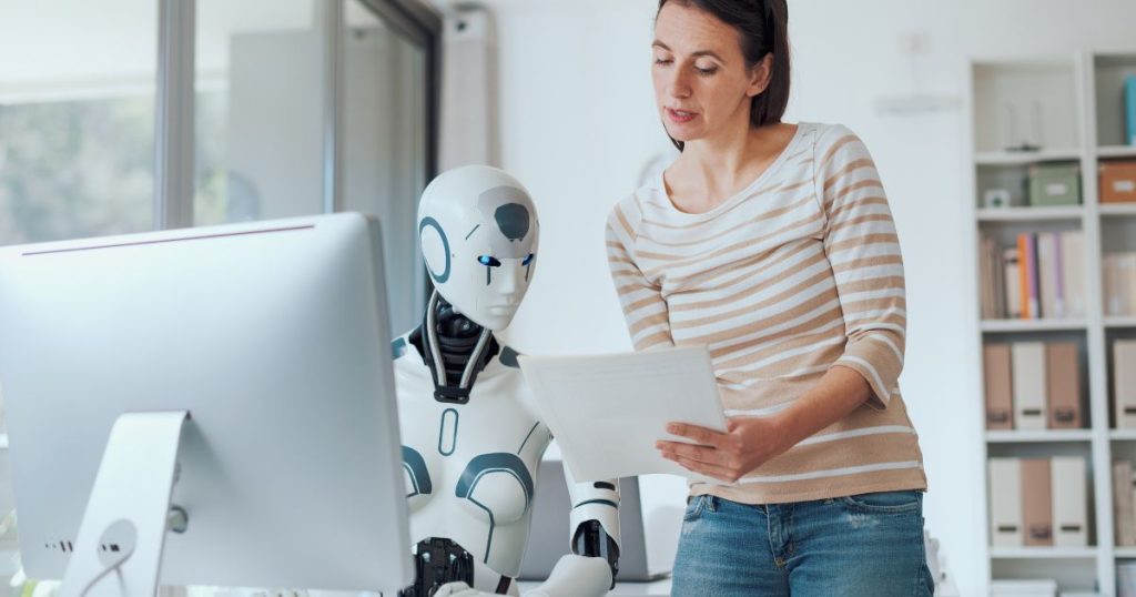 A woman stands next to a humanoid robot, showing it a document as they both look at a computer screen, symbolizing human-robot collaboration.
