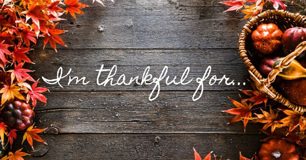 A rustic wooden background with autumn leaves and a basket of seasonal produce surrounds the handwritten phrase “I’m thankful for…”—evoking the spirit of gratitude and Thanksgiving.