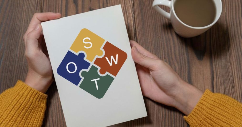 A person wearing a yellow sweater holds a white notebook on a wooden table next to a white coffee mug. The notebook cover displays a colorful SWOT analysis logo shaped like four interlocking puzzle pieces in yellow, red, blue, and green, labeled with the letters S, W, O, and T.